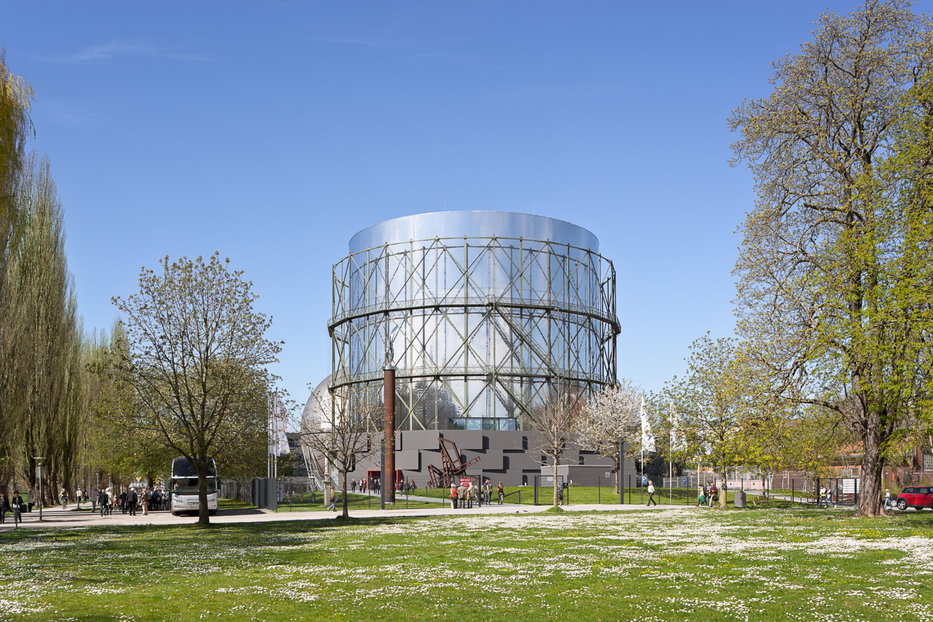 Zylindrischer Gasometer-Rundbau mit Stahlgerüst-Außenhülle, umgeben von Parkanlage mit blühenden Bäumen, Reisebus und Besuchern bei blauem Himmel.