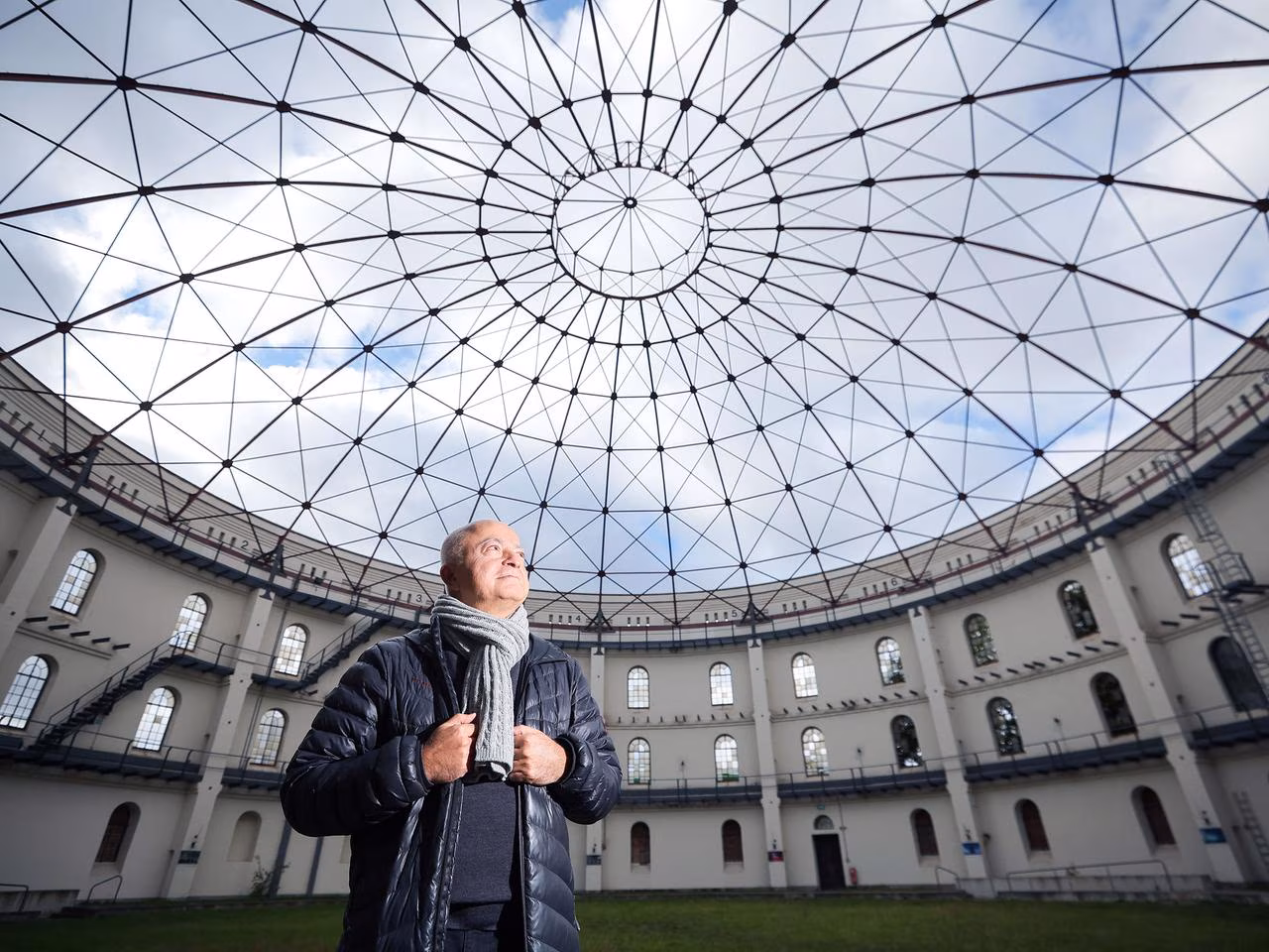 Künstler Yadegar Asisi steht im Innenraum eines Panoramagebäudes unter einer weit gespannten, filigranen Dachkonstruktion.