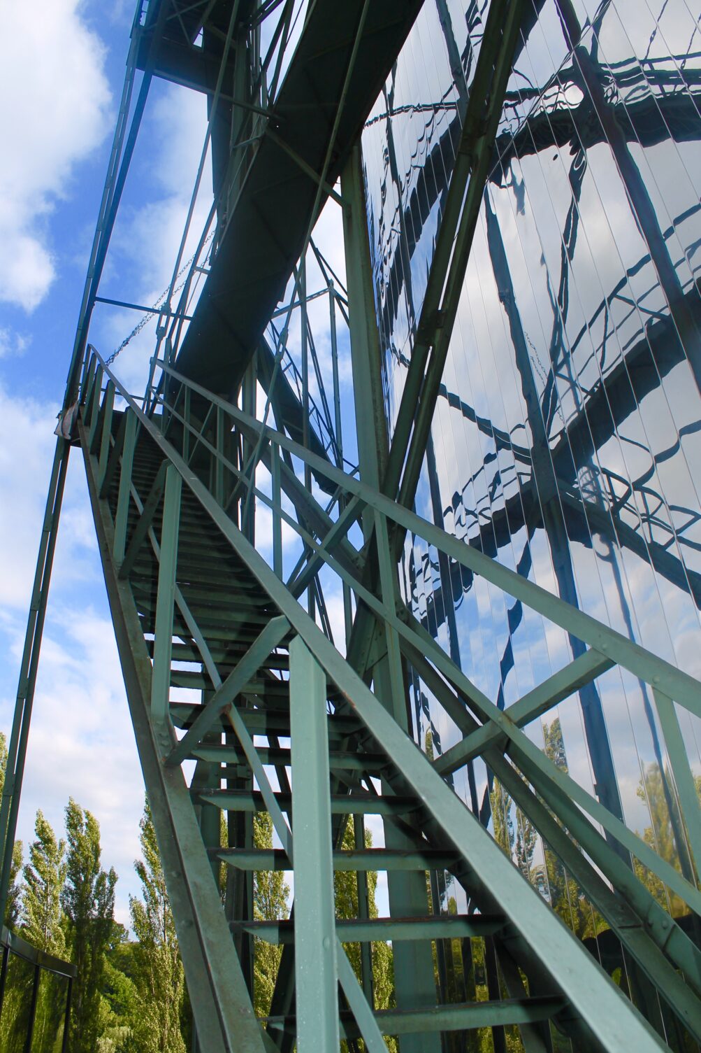Grüne Metalltreppe am Panoramaturm mit spiegelnder Außenwand und Himmel im Hintergrund