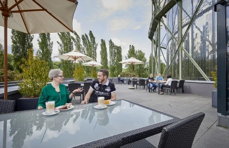Dachterrasse mit Bistrotischen, zwei Personen im Gespräch und Blick auf die Glasfassade des Panoramabaus.